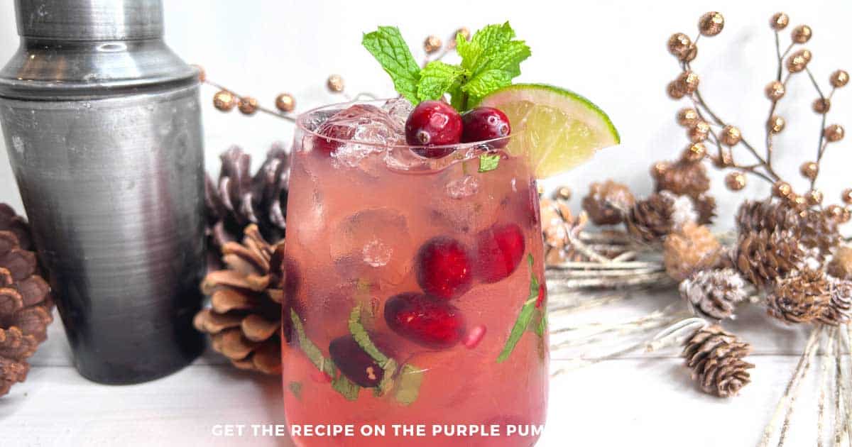 Red cocktail filled with ice, garnished with cranberries, fresh mint and lime. Gold and pinecone Christmas decor is in the background.