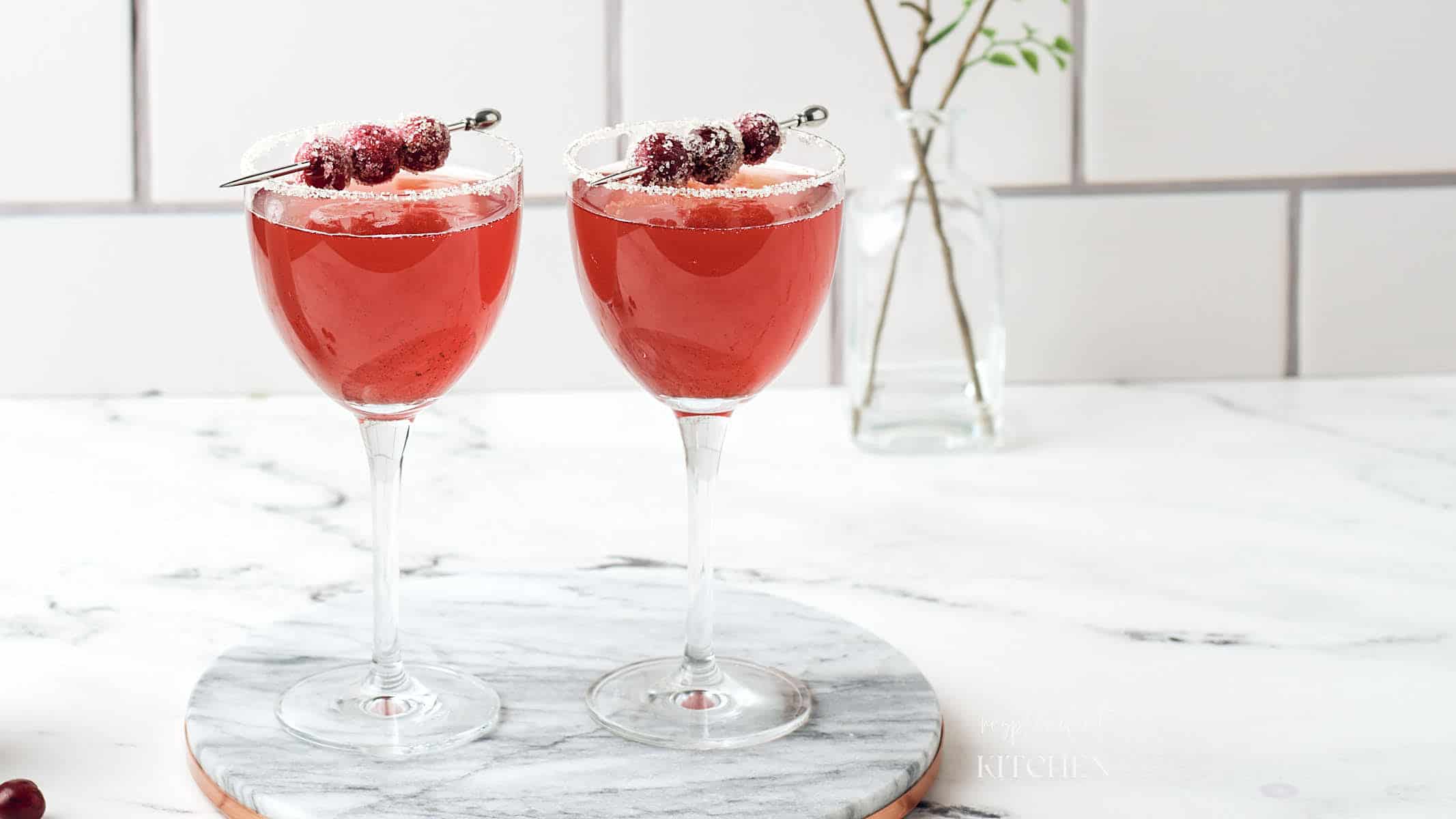 Two stemmed glasses with red drink inside, garnished with a sugared rim and sugared cranberries.