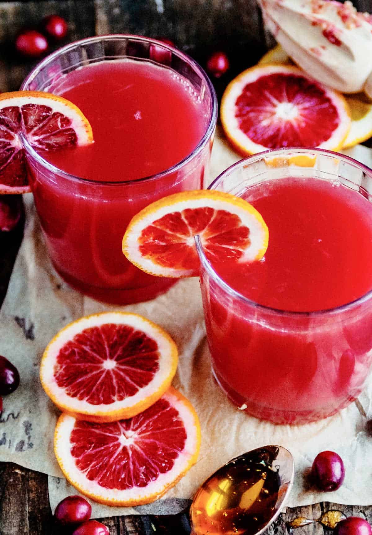 Two vibrant red cocktails garnished with blood oranges on a wooden countertop.