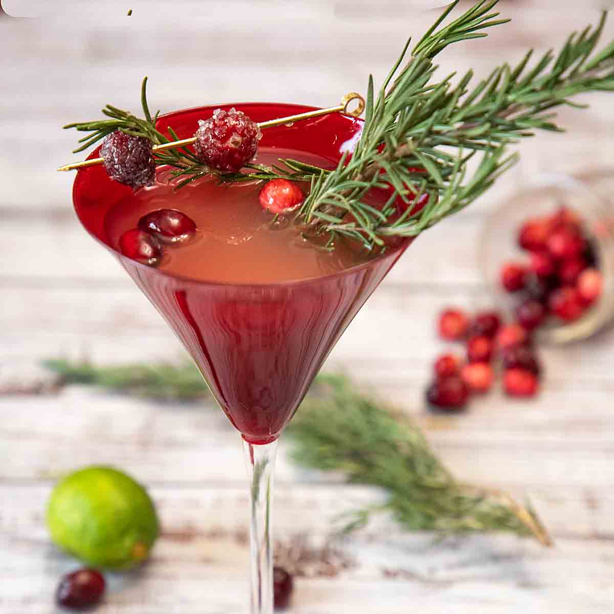 Tall red martini glass filled with a cocktail, and garnished with rosemary and cranberries.