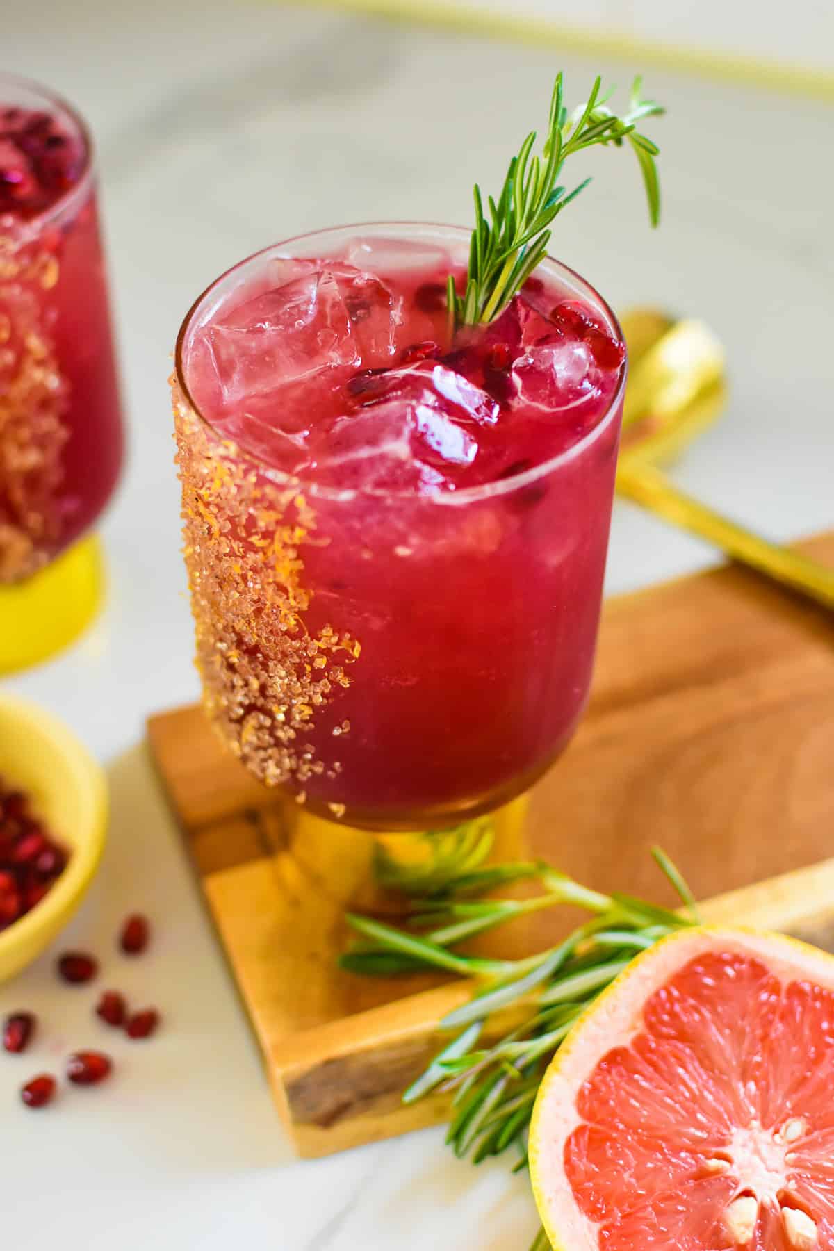 Cocktail glass filled with red liquid and garnished with pomegranate arils, rosemary and a brown sugar coating on the glass.