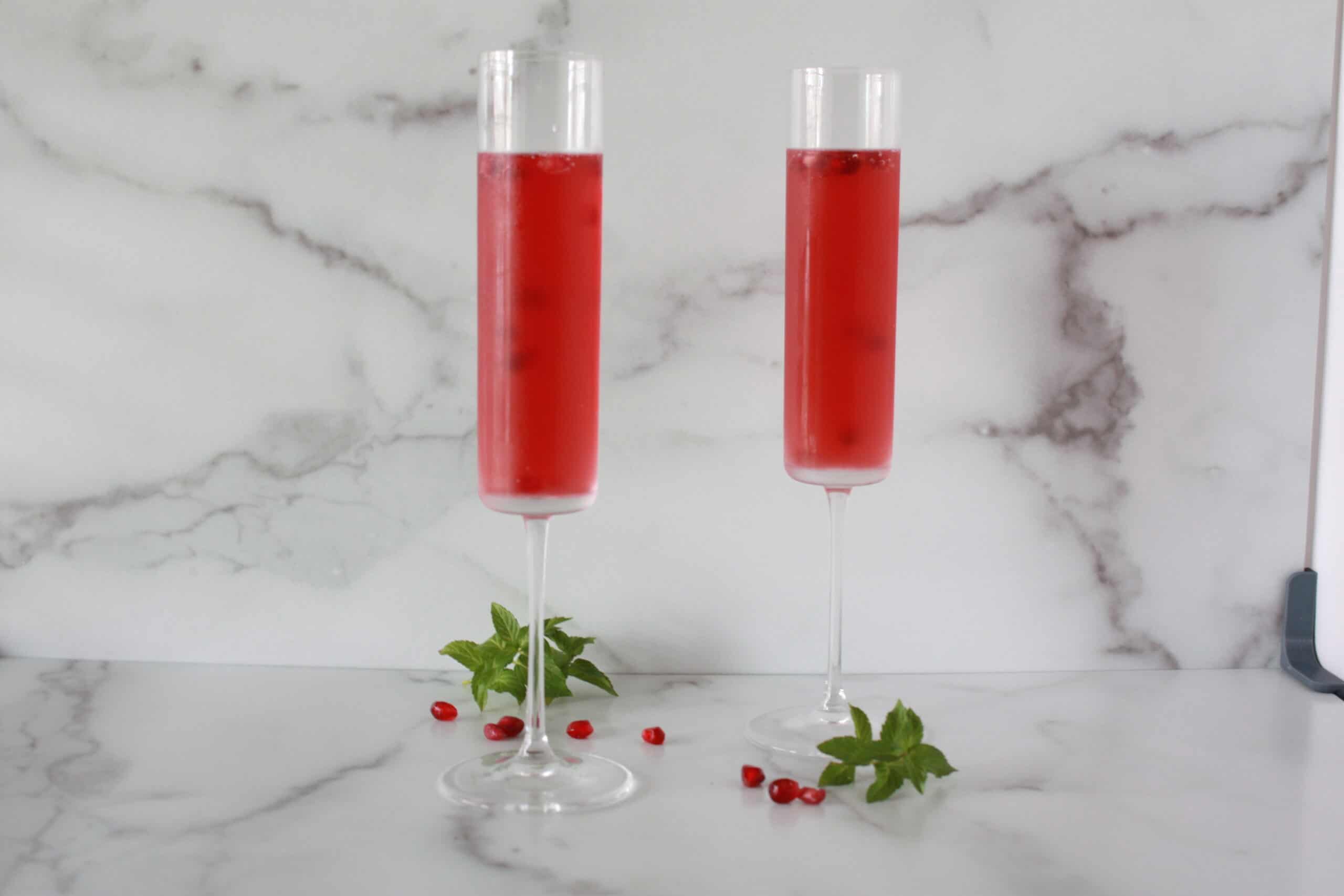 Two tall champagne glasses on a marble countertop filled with red liquid and garnished with pomegranates.