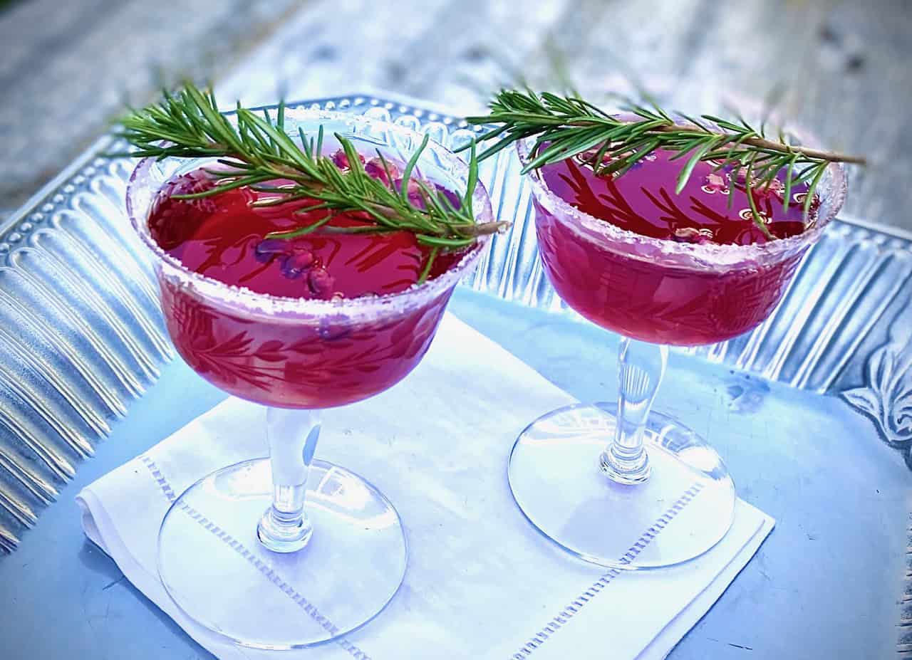 Two cocktail glasses with sugared rims filled with a red cocktail. Garnish is fresh rosemary and pomegranate.