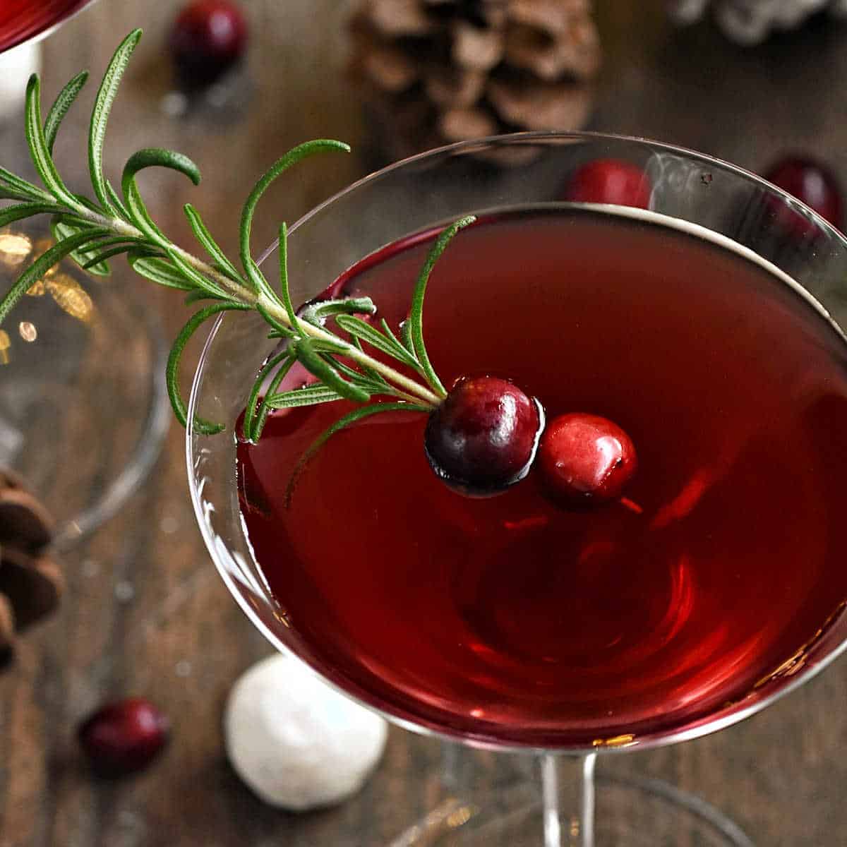 Overhead view of a red colored cocktail in a stemmed martini glass. The drink is garnished with cranberries that are skewered onto a sprig of rosemary.