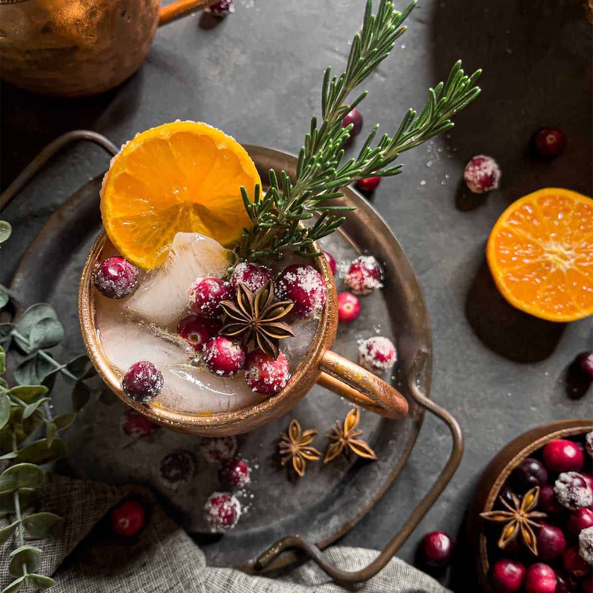 Overhead view of a copper mug with a cocktail in it, garnished with star anise, sugared cranberries, an orange slice and fresh rosemary.