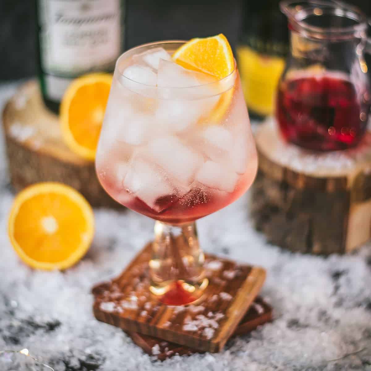 A stemmed cocktail glass filled with ice and a bubbly cocktail, garnished with an orange slice. Winter decor is all around the drink.