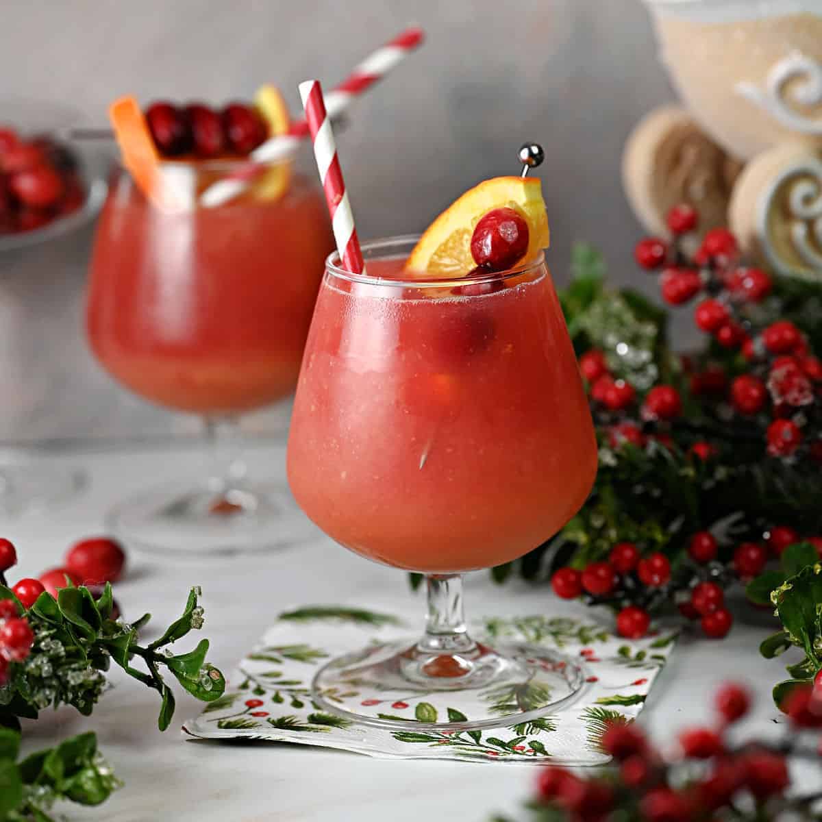 A cocktail glass filled with an orangey red mixture with an orange slice, cranberries and a red and white straw sits on a countertop on top of a Christmas napkin. Christmas decor is all around and there is another of the same drink in the background.