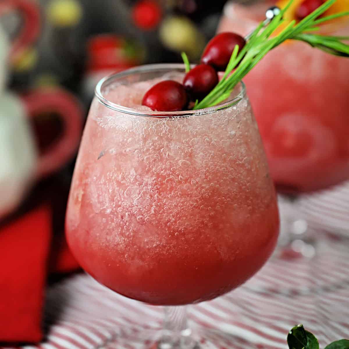 Red colored frozen cocktail in a wide goblet style glass. Rosemary and cranberries garnish the drink on the top, and there is Christmasy decor around the glass.