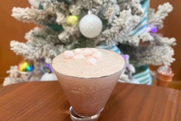 A light brown colored, creamy frozen cocktail in a martini glass on a wooden table, garnished with mini marshmallows. A Christmas tree is in the background.