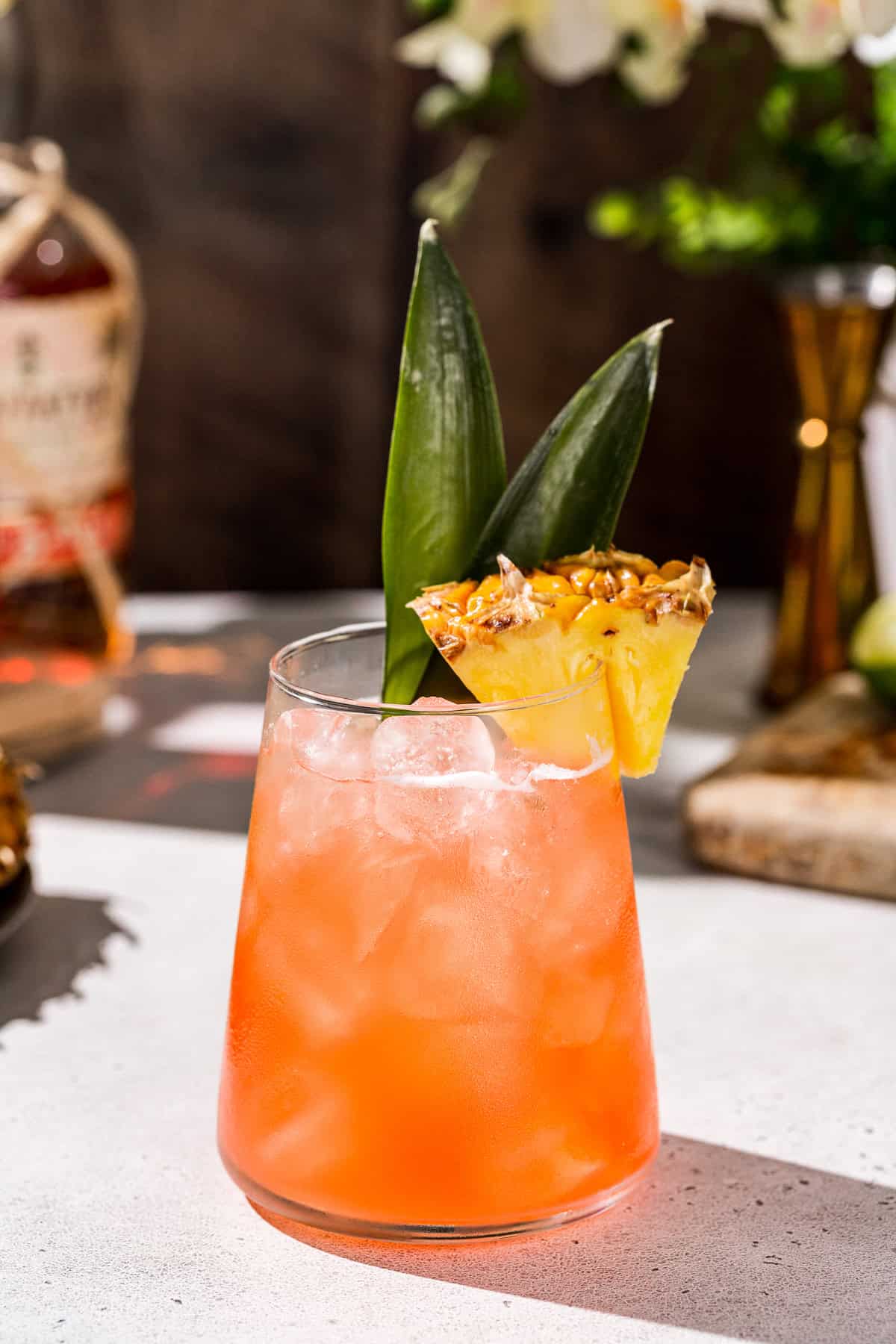 Side view of a Jungle Bird cocktail, served over ice with a pineapple wedge and pineapple fronds for garnish. In the background are a bottle of rum, a gold jigger and tropical decor.