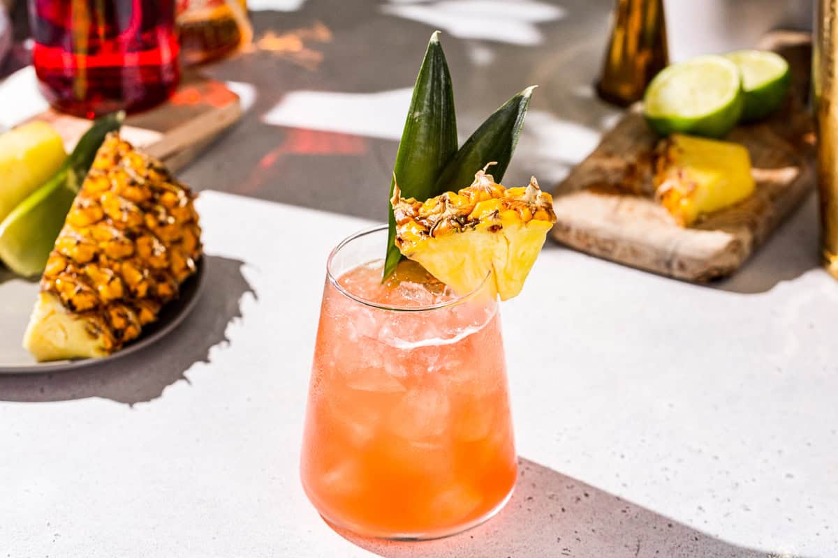 Slightly overhead view of an orange colored Jungle Bird cocktail, garnished with a piheapple piece and pineapple fronds.