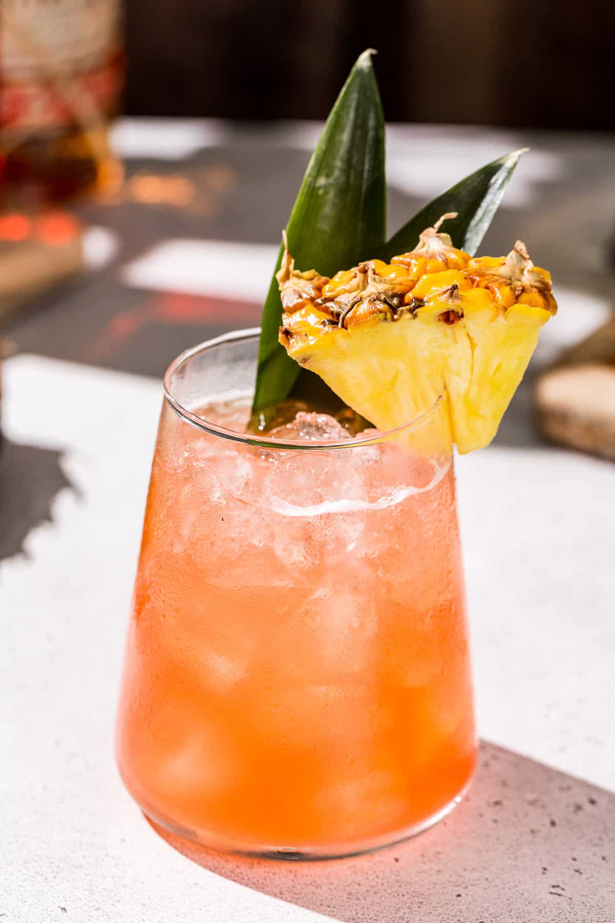 Close up of a Jungle Bird cocktail on a countertop. The drink is reddish orange and is garnished with a pineapple wedge and pineapple fronds.