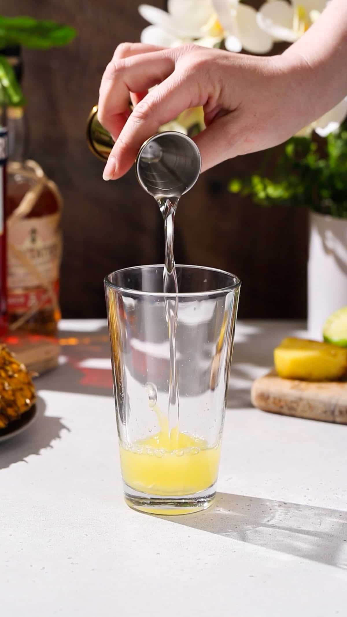 Hand adding simple syrup to a cocktail shaker filled with yellow liquid.