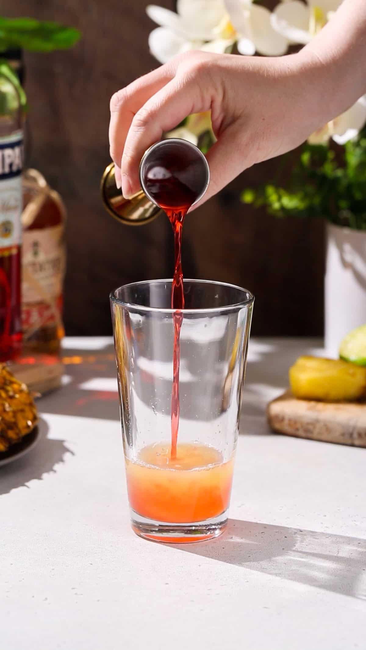 Hand adding campari liqueur to a cocktail shaker filled with yellow liquid.