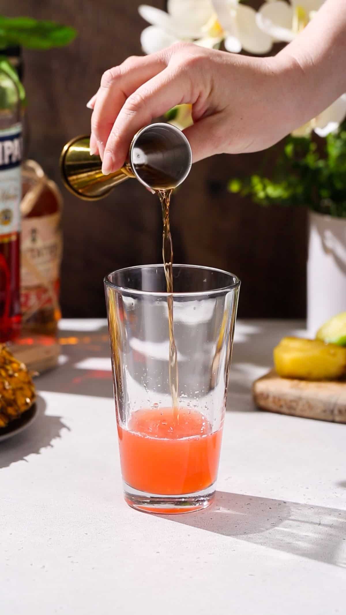 Hand adding dark pot still rum to a cocktail shaker filled with red liquid.