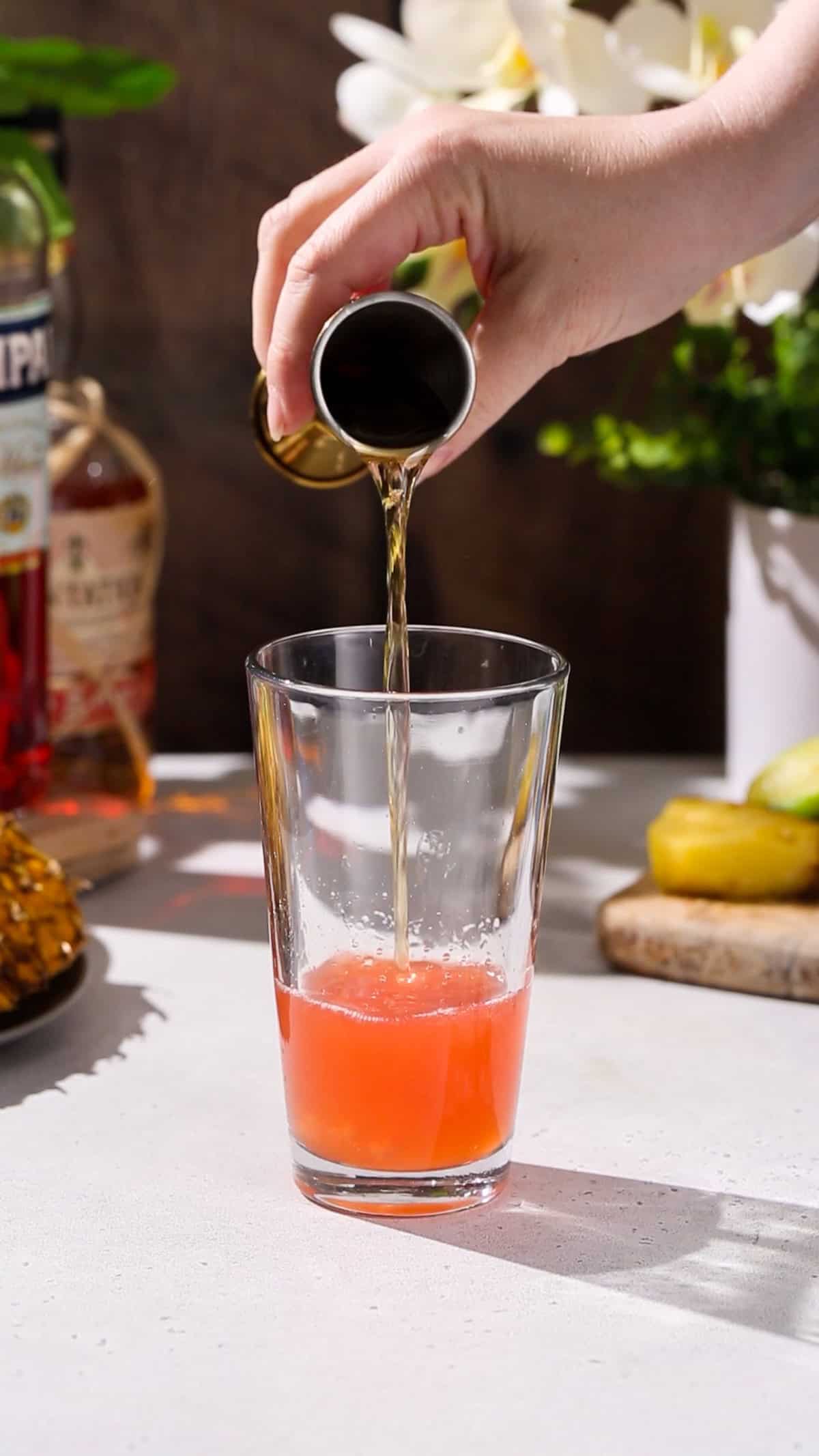 Hand adding dark rum to a cocktail shaker filled with red liquid.