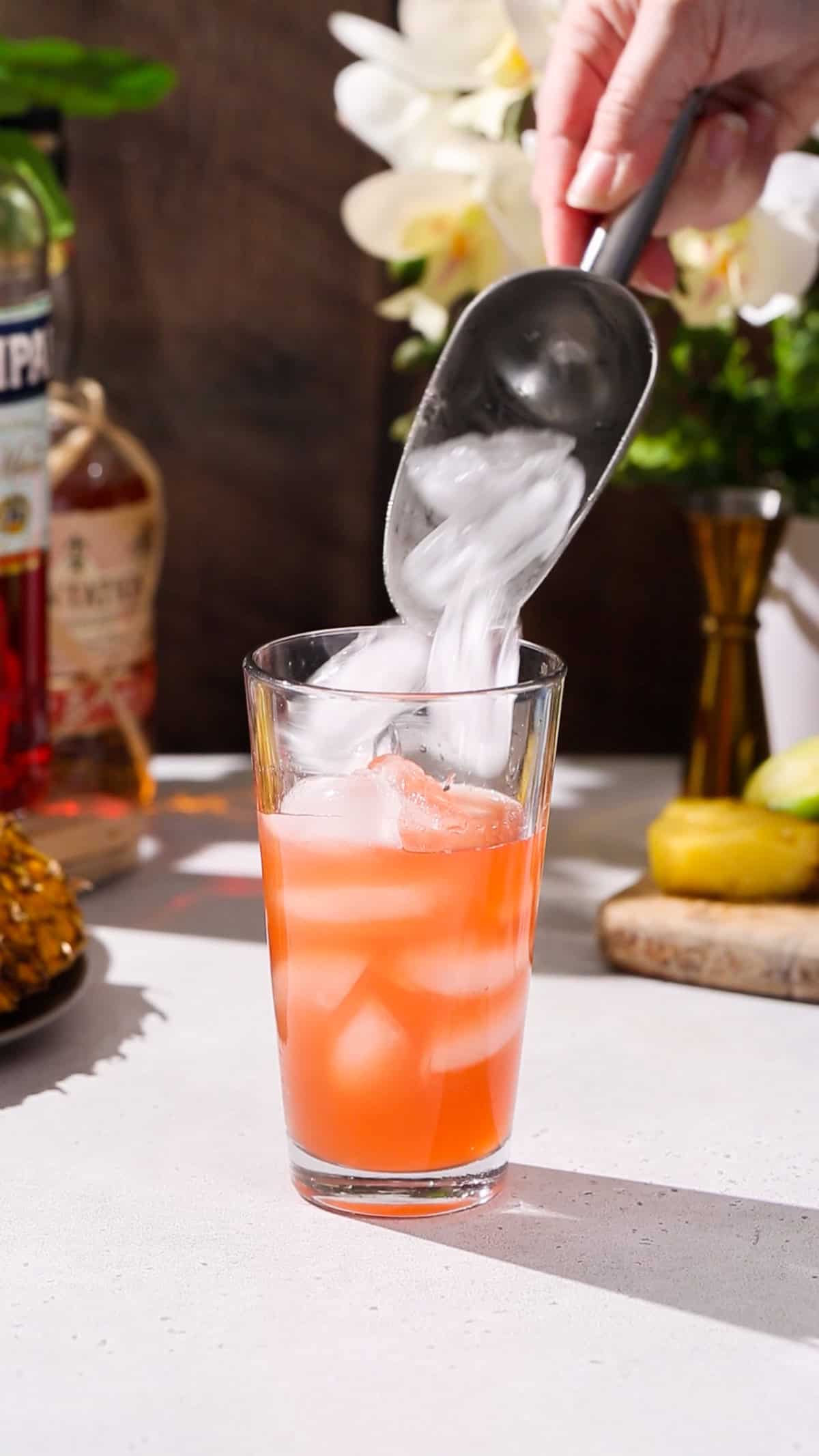 Hand using an ice scoop to add ice to a glass cocktail shaker filled with orange liquid.