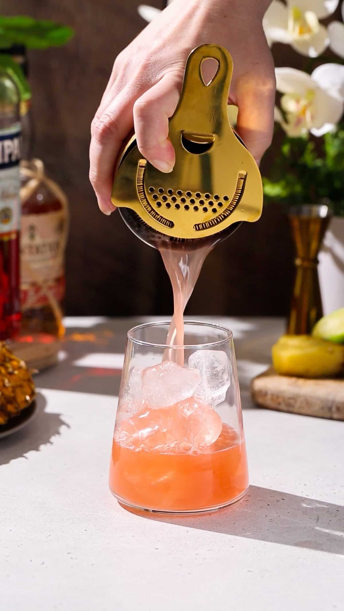 Hand using a cocktail shaker to strain an orange colored cocktail into a glass filled with ice.