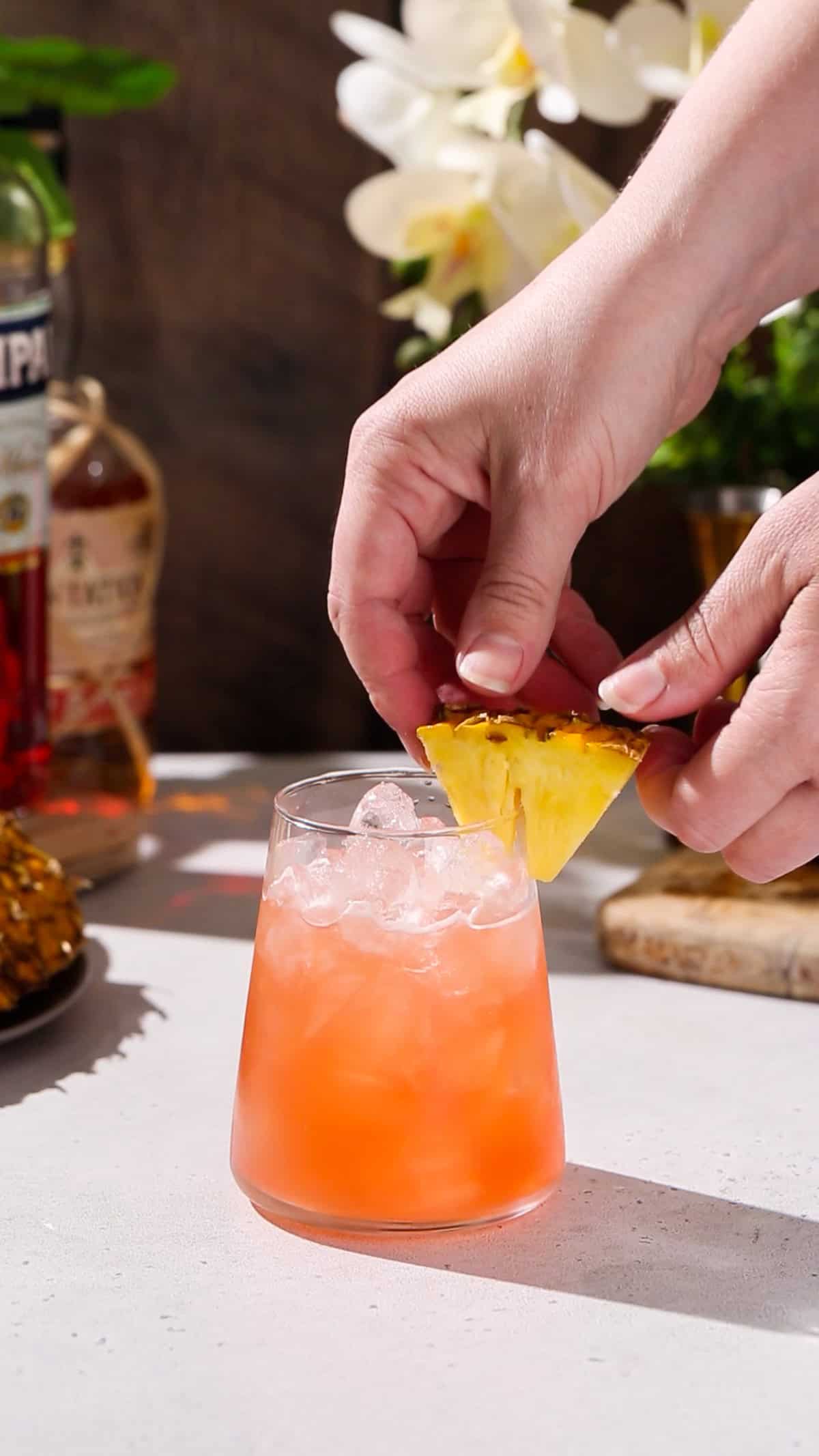 Hand adding a wedge of pineapple to the rim of a cocktail glass filled with reddish orange liquid.