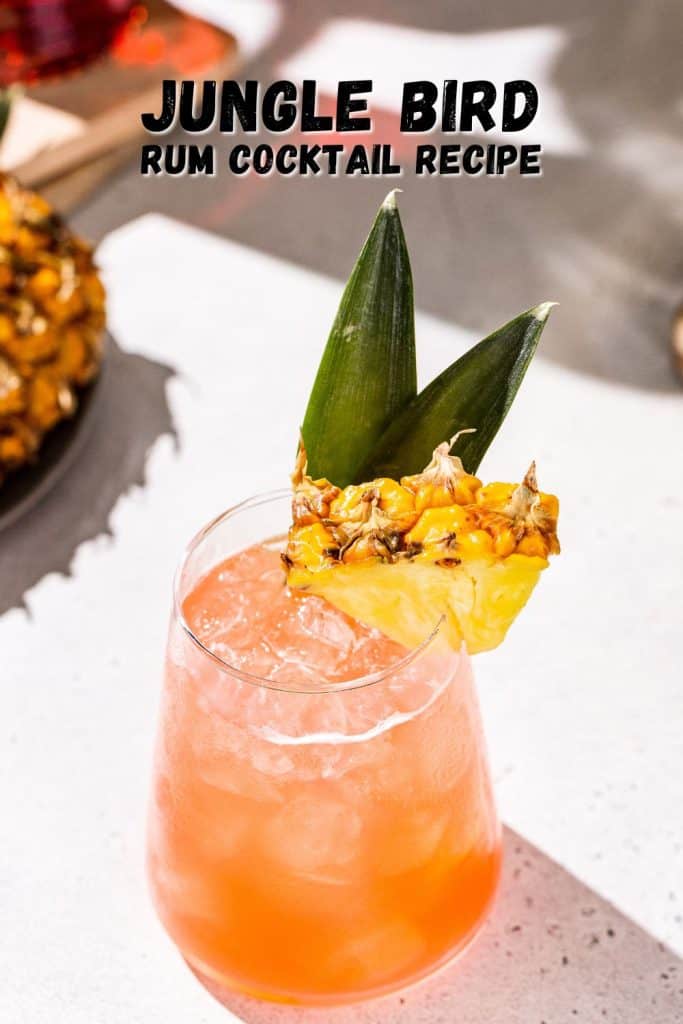 Overhead view of a Jungle Bird Cocktail in a stemless wine glass. The drink is reddish orange and served on ice. The glass is garnished with a pineapple wedge and two pineapple fronds. Text overlay above the drink says “Jungle Bird rum cocktail recipe” in black lettering.