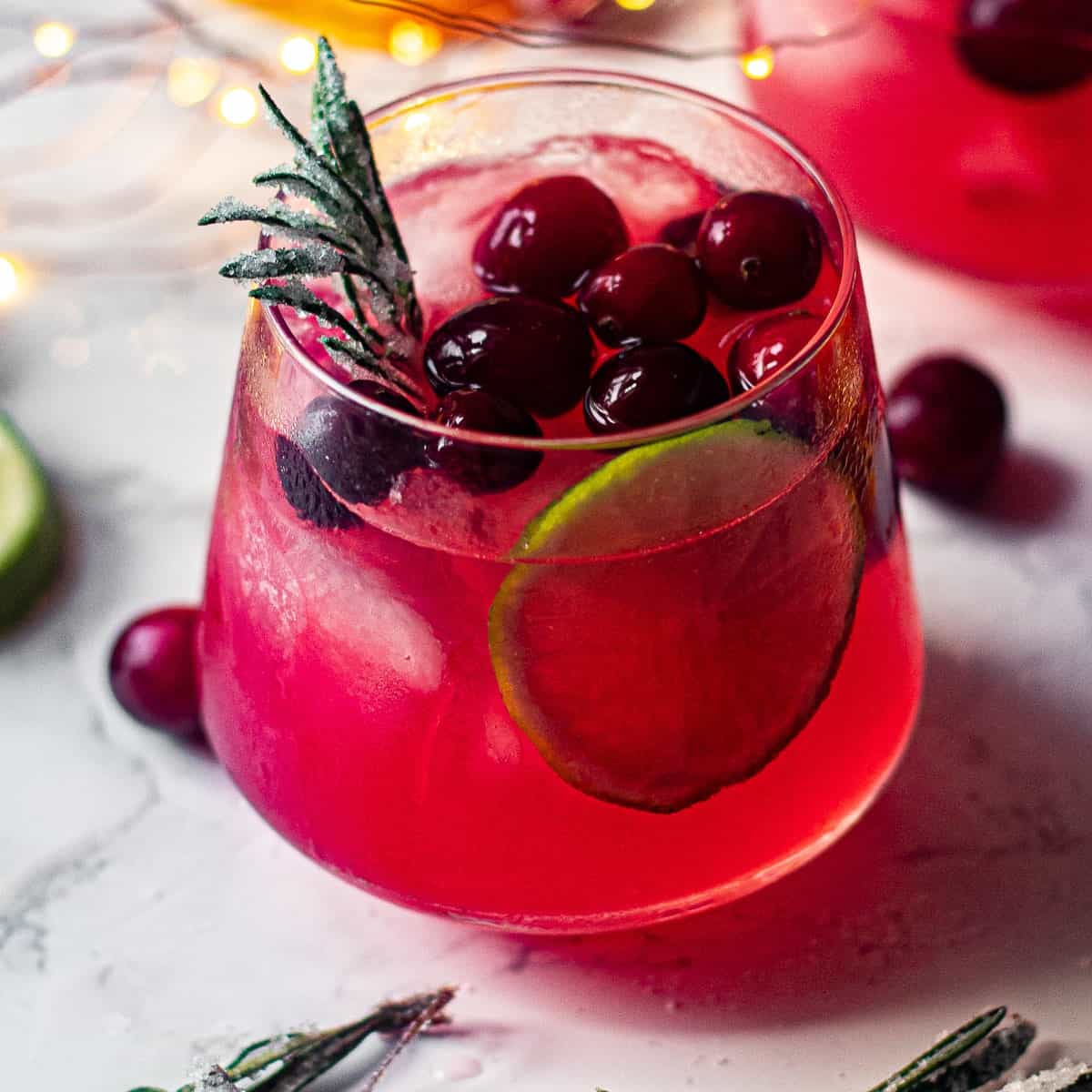 Short cocktail glass filled with red liquid, garnished with cranberries, a lime slice and sugared rosemary sprig.