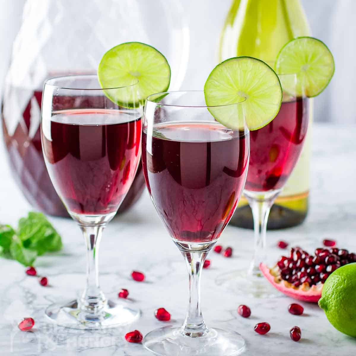 Three small goblets each filled with a dark red cocktail and garnished with a lime slice. Fresh pomegranate seeds are all around on the countertop.