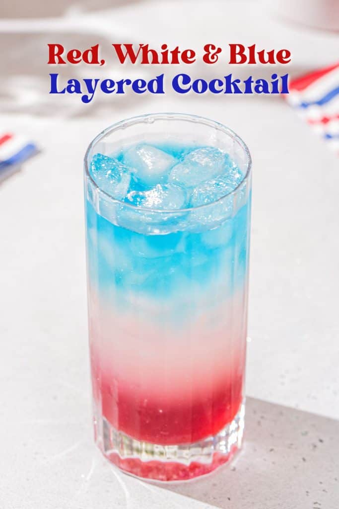 Layered cocktail in a tall collins glass on a countertop. The drink has a blue layer on top, a white layer in the middle and a red layer on the bottom. Text above the drink says “Red, White & Blue Layered Cocktail”.