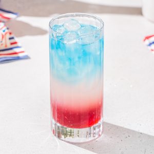 Red, white and blue layered cocktail in a tall glass sitting on a countertop. In the background are some striped red, white and blue napkins.