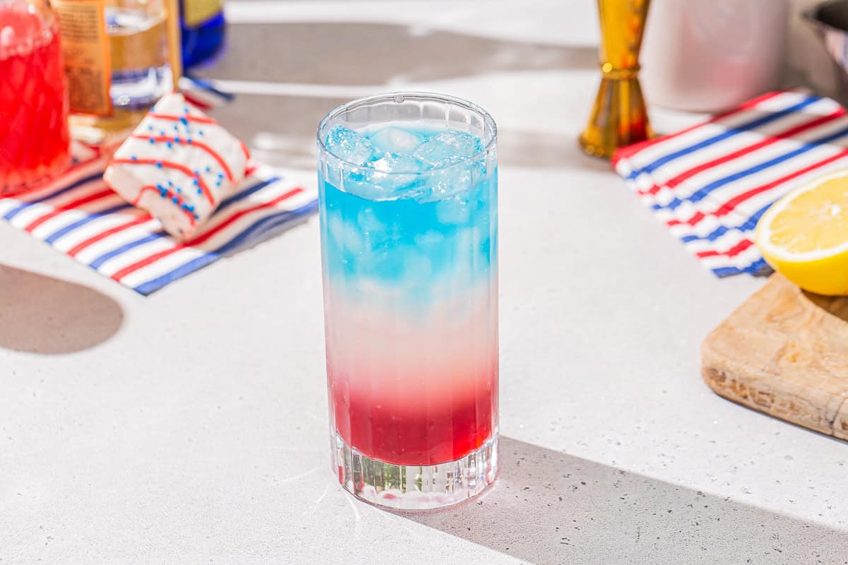 Red, white and blue cocktail with the blue layer on top sitting on a countertop. In the background are some striped napkins and red, white and blue cakes.