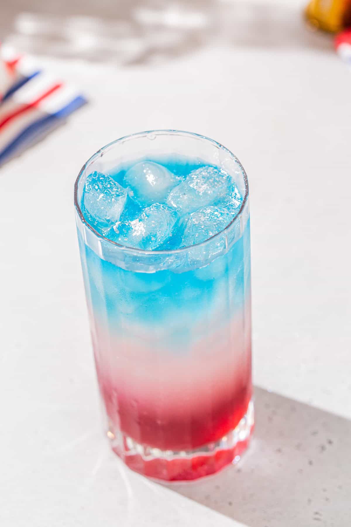 Close up view of a layered cocktail with a blue layer on top, white in the middle and red on the bottom. The drink is in a tall Collins glass.