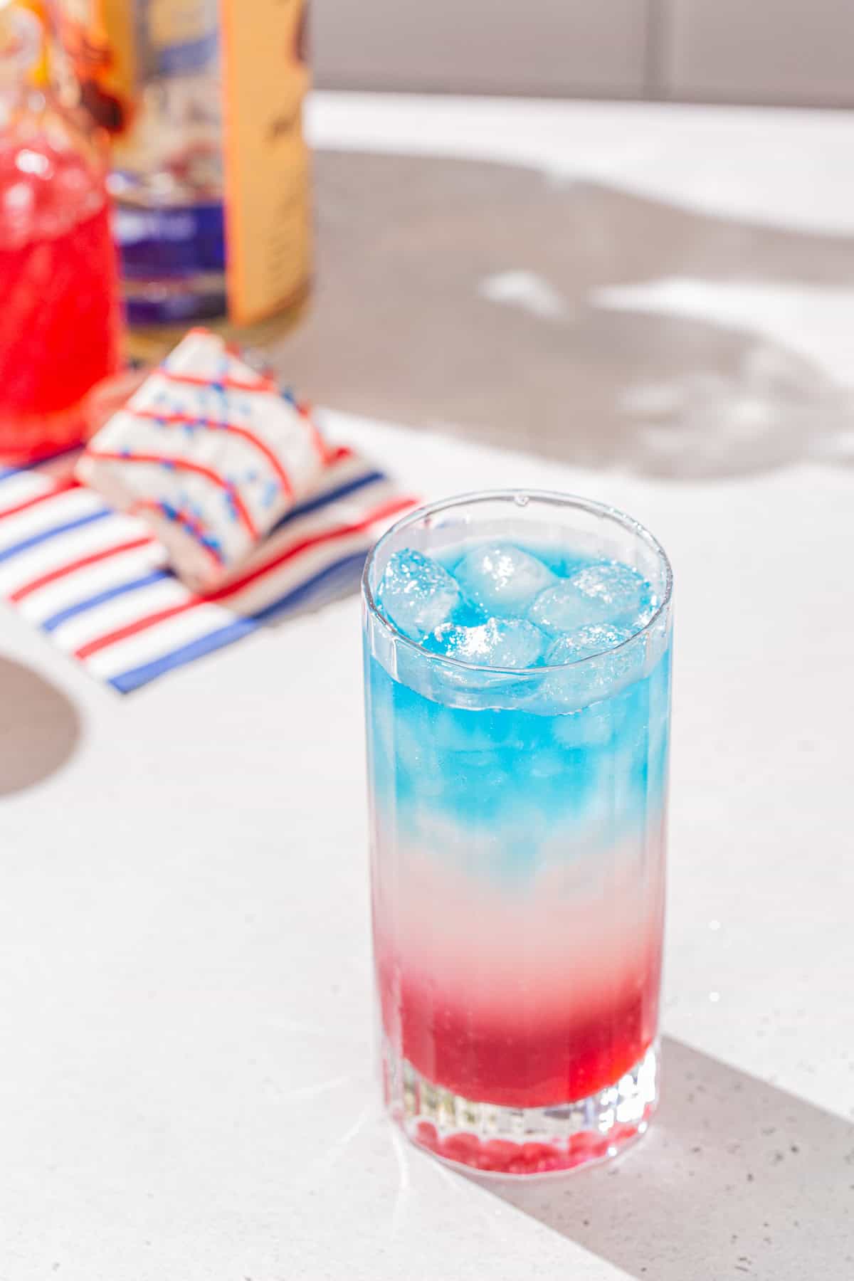 A red, white and blue layered cocktail on a countertop with a bottle of red syrup, vodka, blue Curacao and some red, white and blue decor in the background.