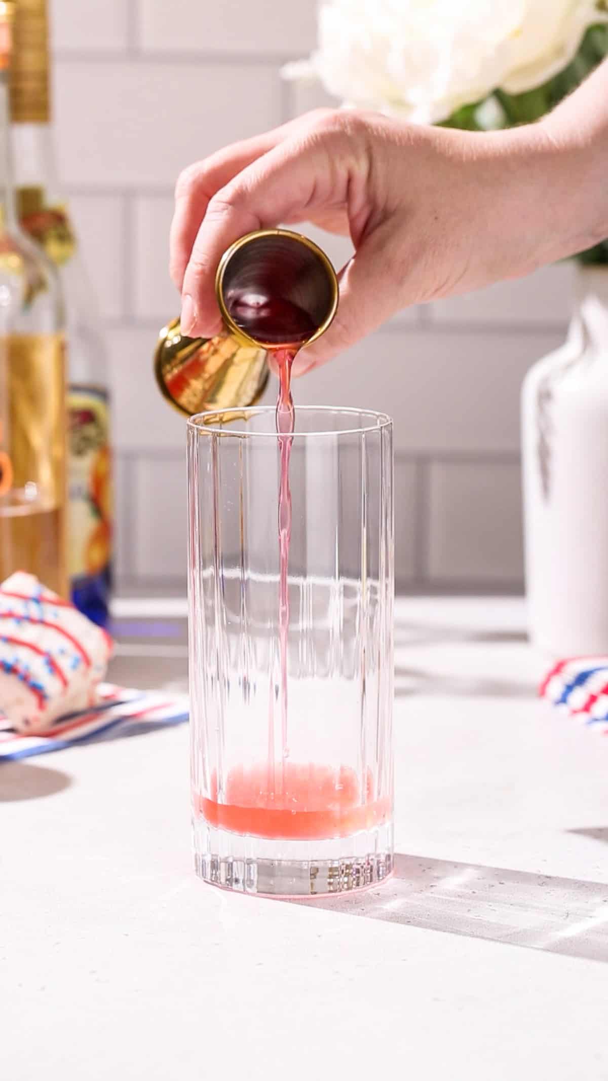 Hand pouring raspberry syrup into a tall cocktail glass.