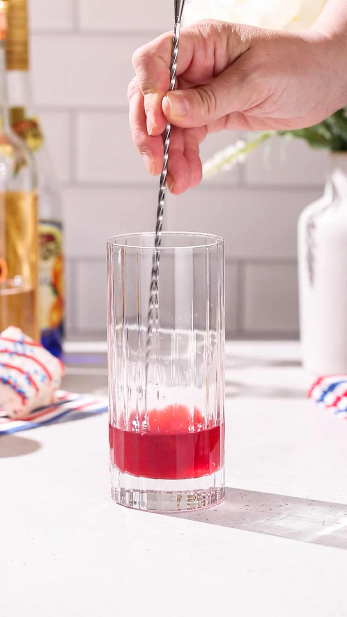 Hand stirring red liquid in a collins glass with a long bar spoon.