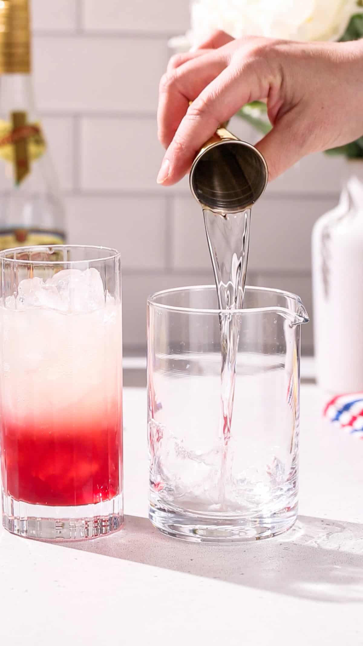 Hand pouring vodka into a cocktail mixing glass. Off to the left is a cocktail glass with a red and white layer.