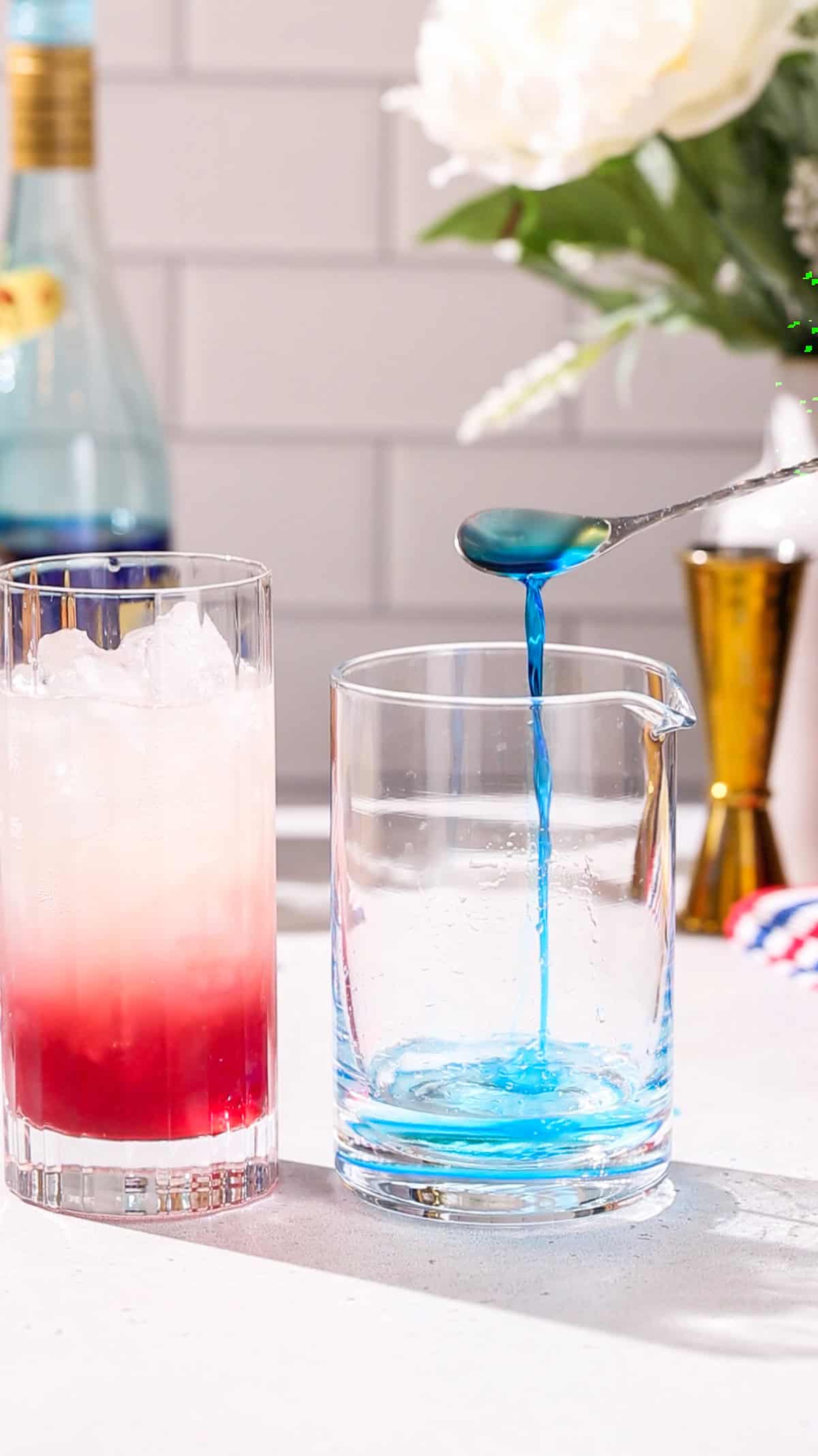 Hand using a bar spoon to add blue curacao liqueur to a cocktail mixing glass. Off to the left is a red and white cocktail in a glass.