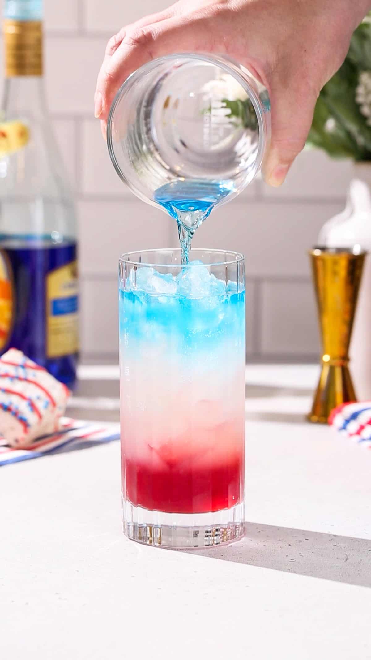 Hand pouring blue liquid into a cocktail glass with a white and red layer in it.