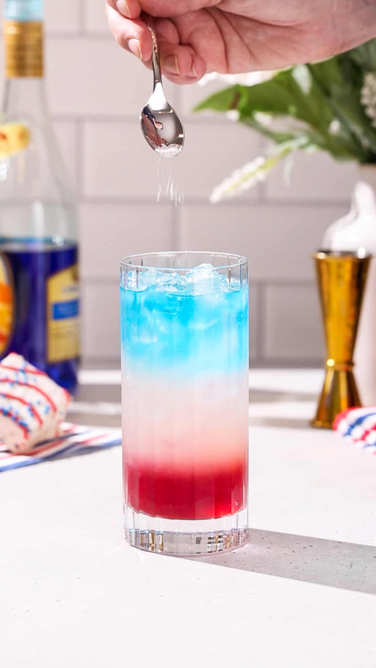 Hand using a spoon to add sparkling glitter to the top of a red, white and blue cocktail.