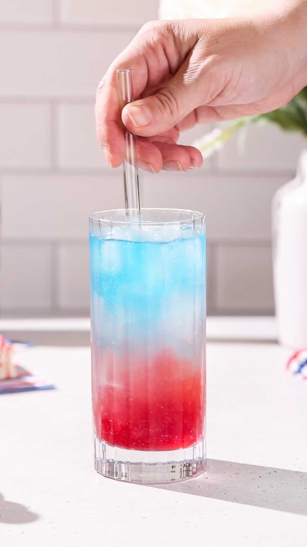 Hand stirring a red, white and blue cocktail with a clear straw.