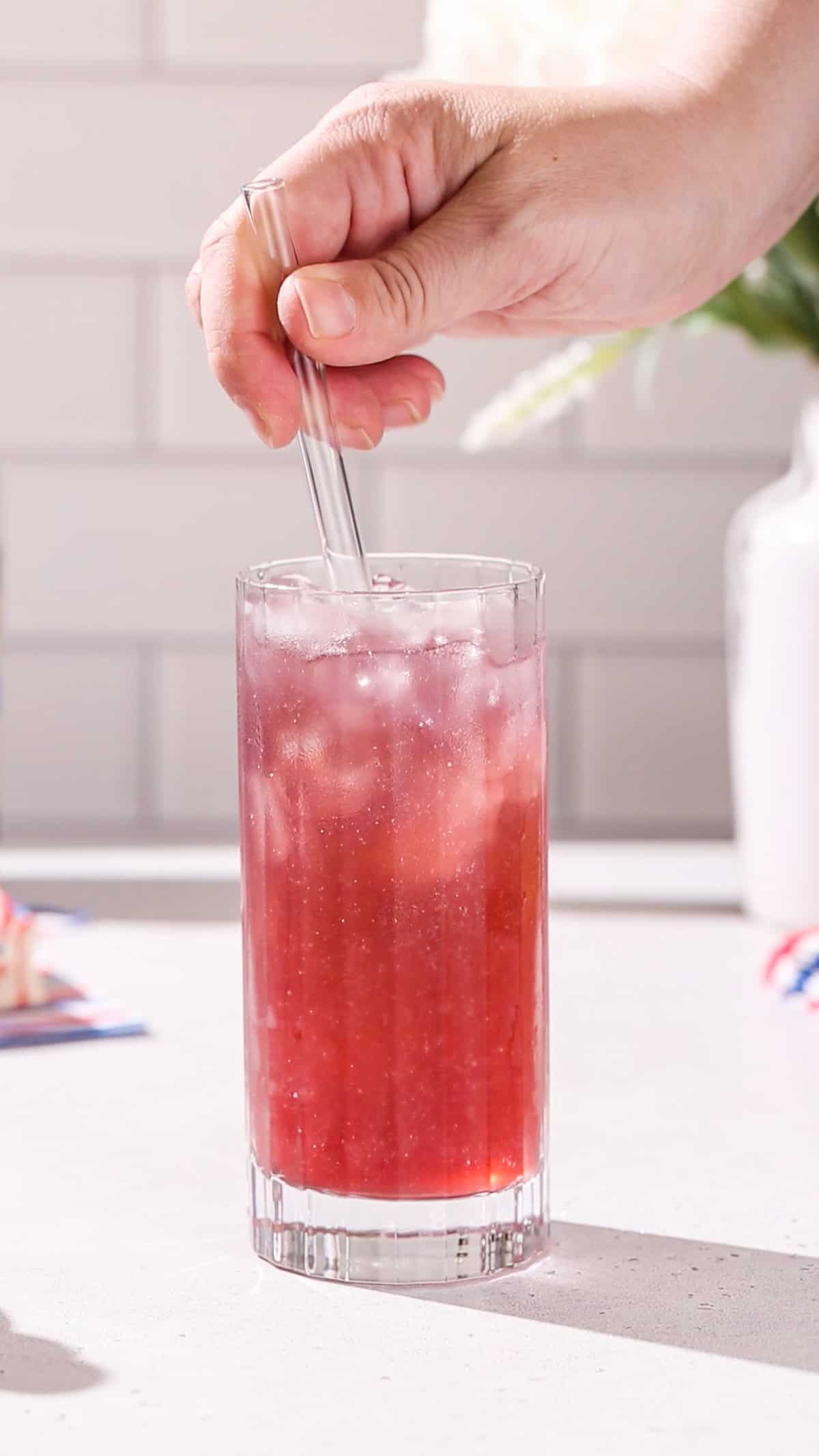 Hand stirring a red cocktail with a clear straw.