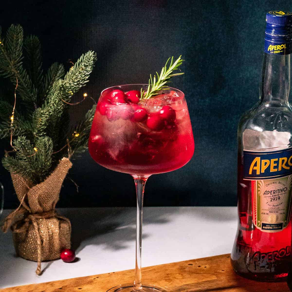 Tall stemmed spritz glass filled with red liquid and garnished with cranberries and rosemary sprig. A bottle of Aperol is off to the right and a mini Christmas tree to the left.