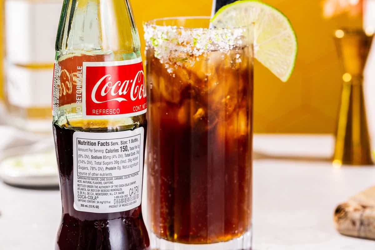 Close up of the ingredients label of a Mexican Coca-Cola bottle. A Batanga cocktail is in the background.