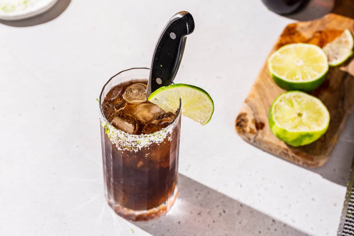 Overhead view of a Batanga cocktail garnished with a knife along with a lime slice and salted rim. A cutting board with a cut lime is in the background.