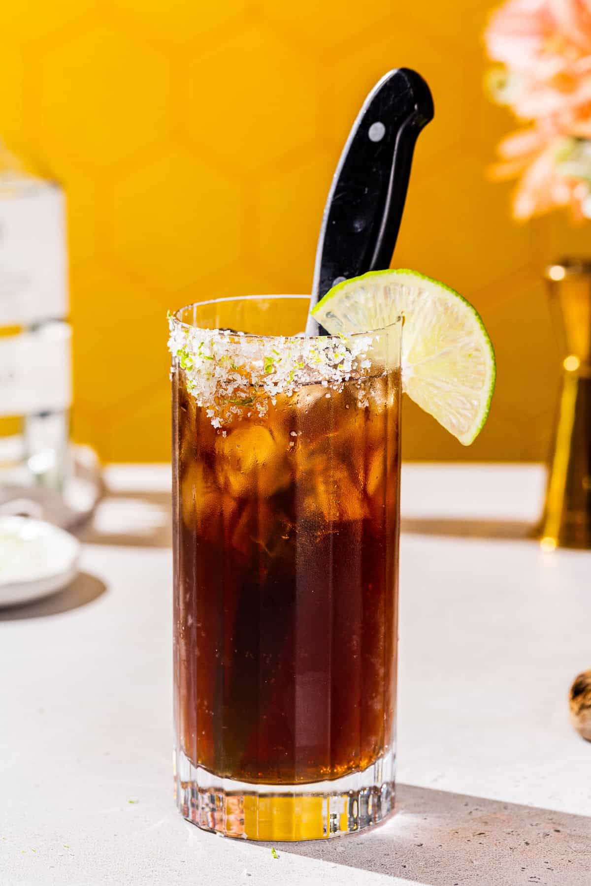 Side view of a Batanga cocktail with a bottle of tequila and jigger in the background.