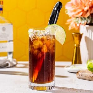 Side view of a Batanga cocktail on a countertop with ingredients used to make the drink in the background.