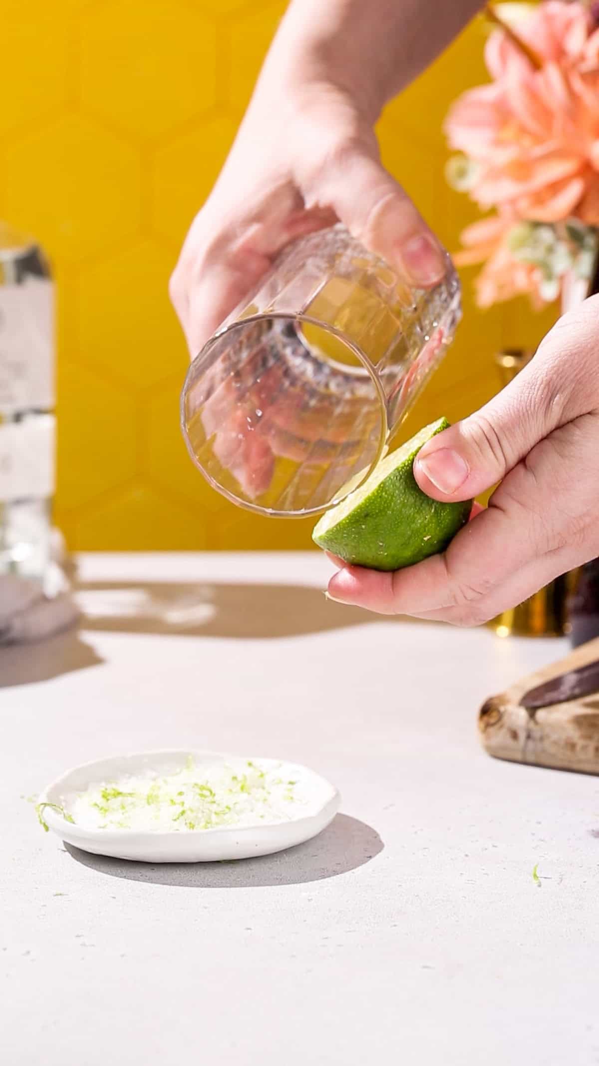 Hand using a cut lime to wet the rim of a tall cocktail glass.