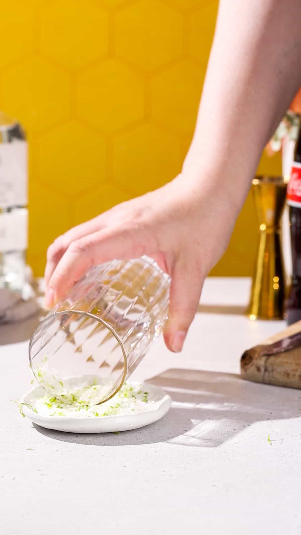 Hand dipping the rim of a tall cocktail glass into a dish of lime salt.