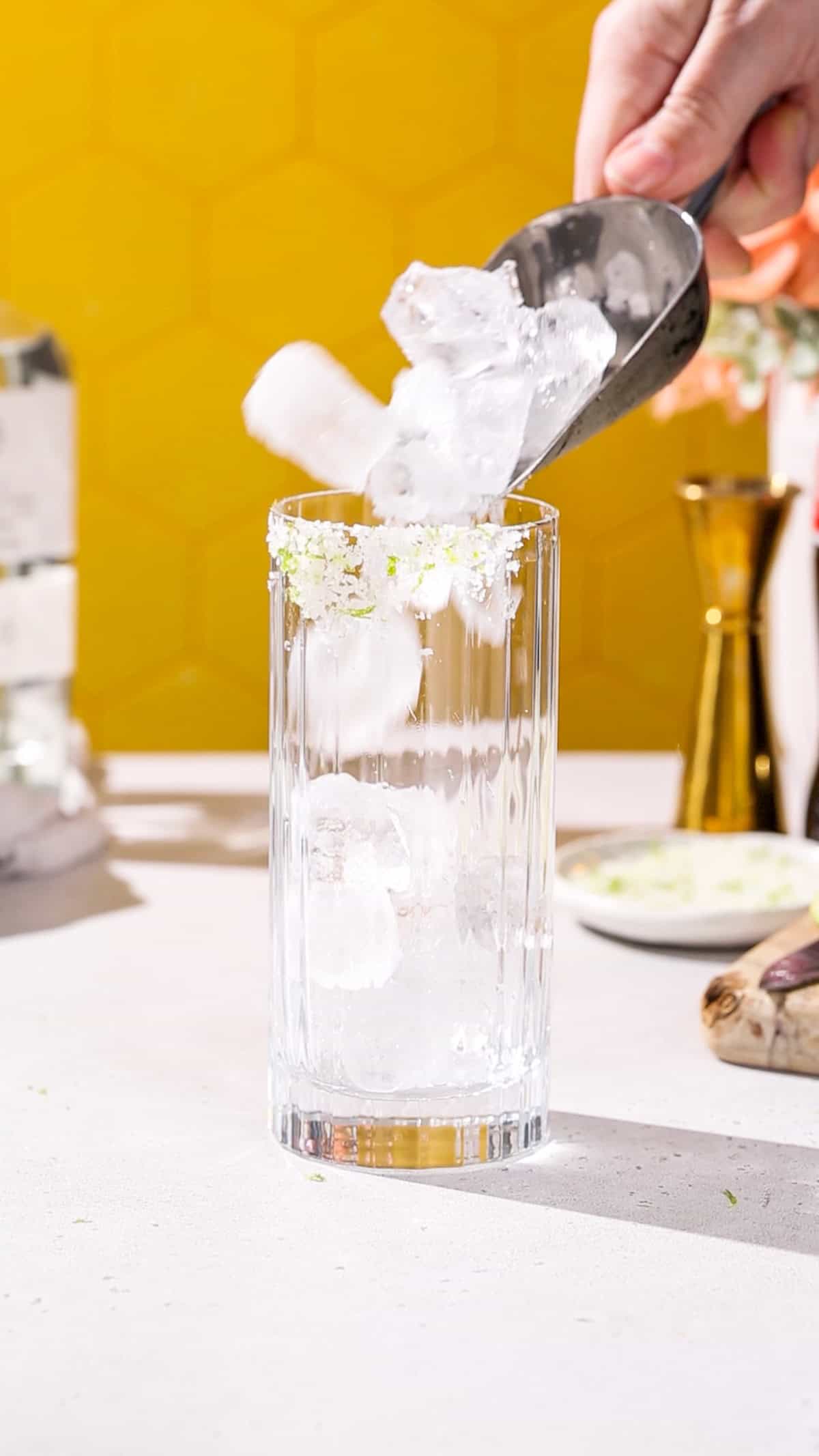 Hand using an ice scoop to fill a tall cocktail glass with ice.