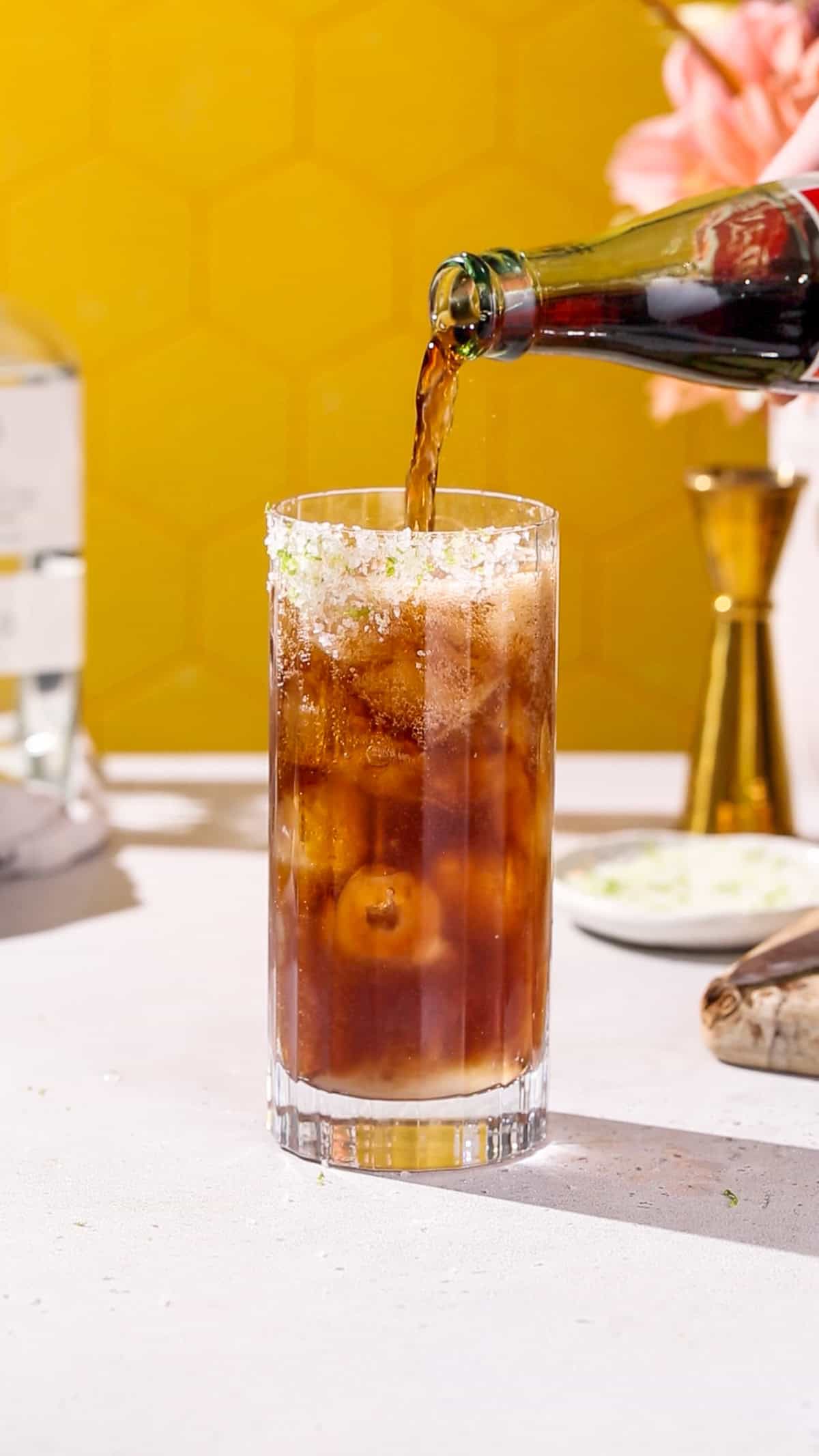 Hand pouring Mexican Coke into a cocktail glass filled with ice and with a lime salt rim.