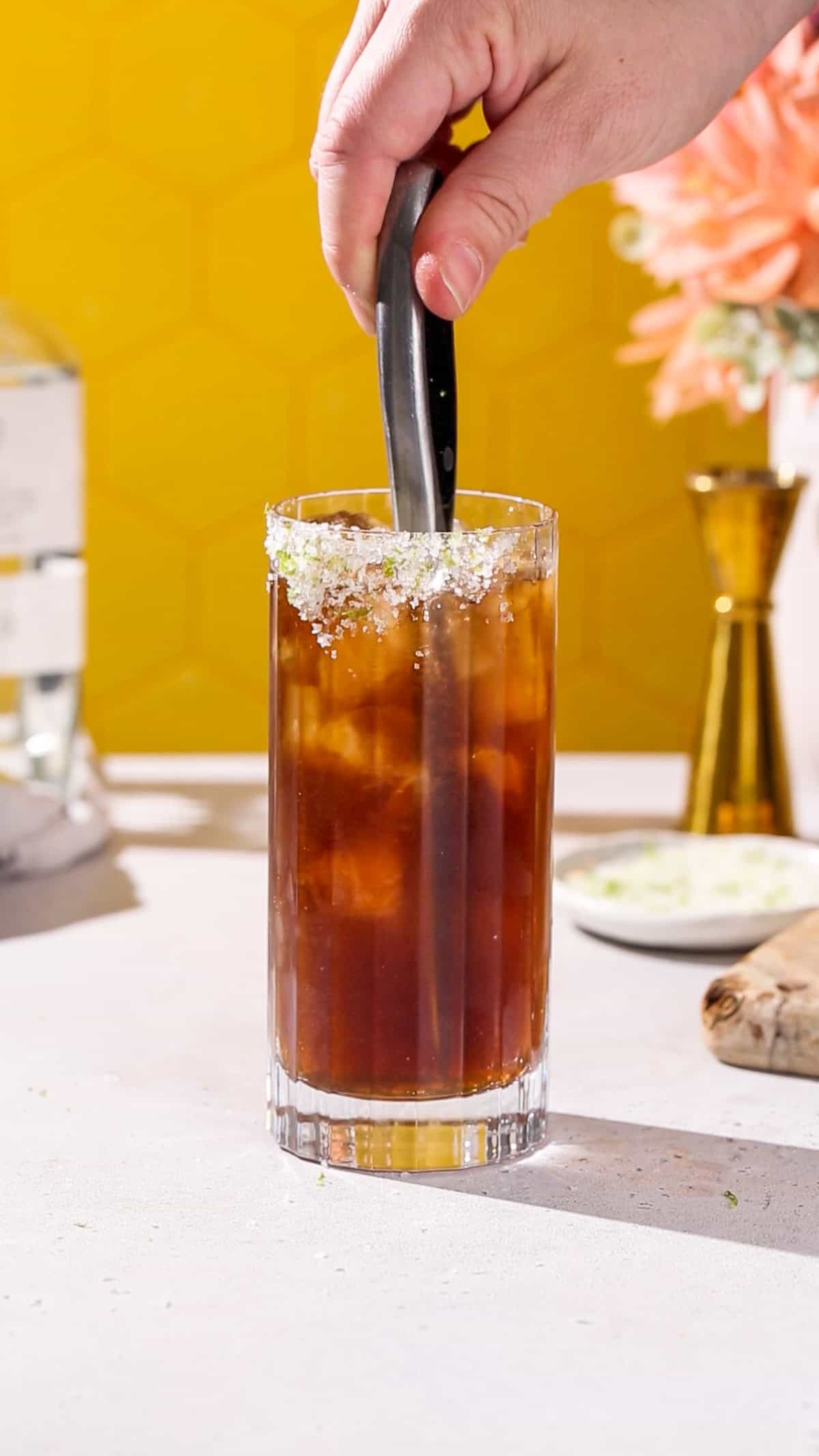 Hand using a knife to stir a dark colored cocktail in a tall glass.