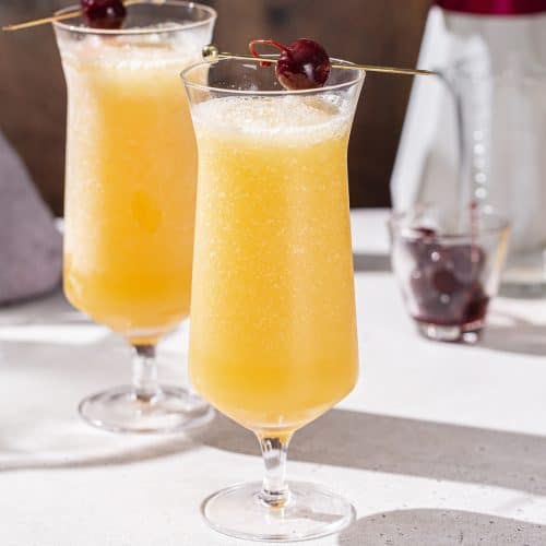 Side view of hurricane glasses filled with frozen whiskey sours. The drinks are yellow in color and have cocktail cherries on top.